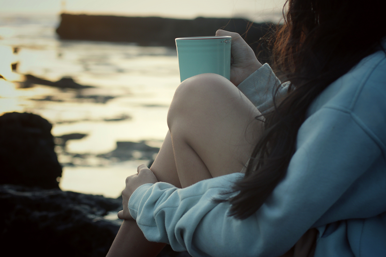 Cultivating Self-care for Sustained Change a young woman in comfy clothes drinking tea on a rocky shore