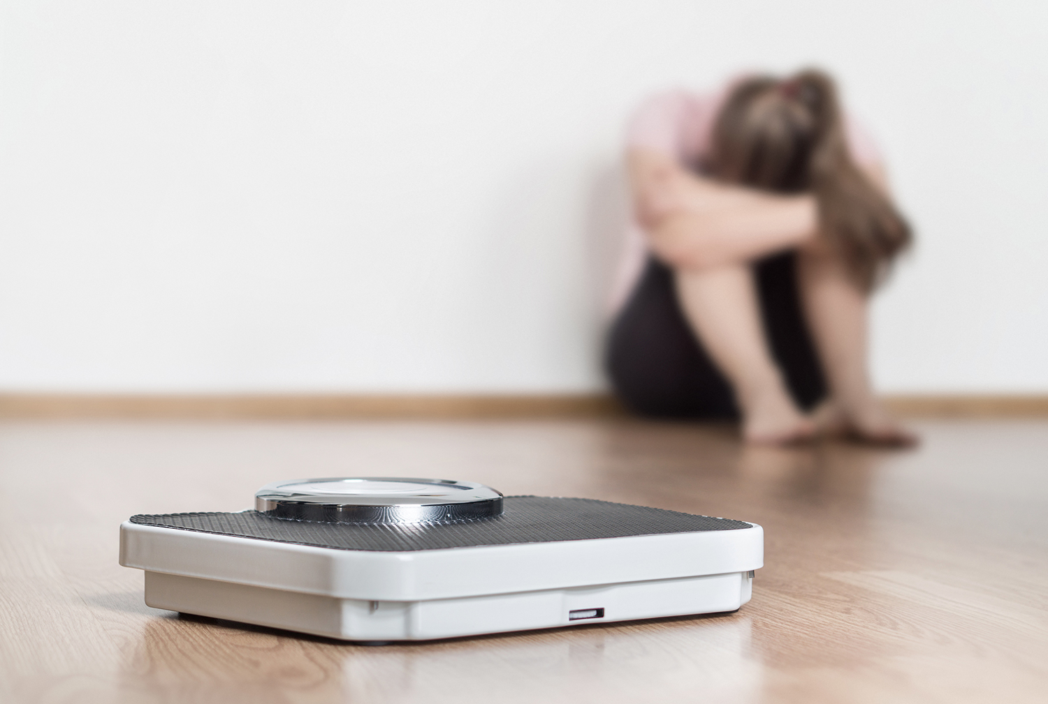 Reasons Why You Can’t Lose Weight A scale on the floor with a woman sitting in the background with her head hung with failure.