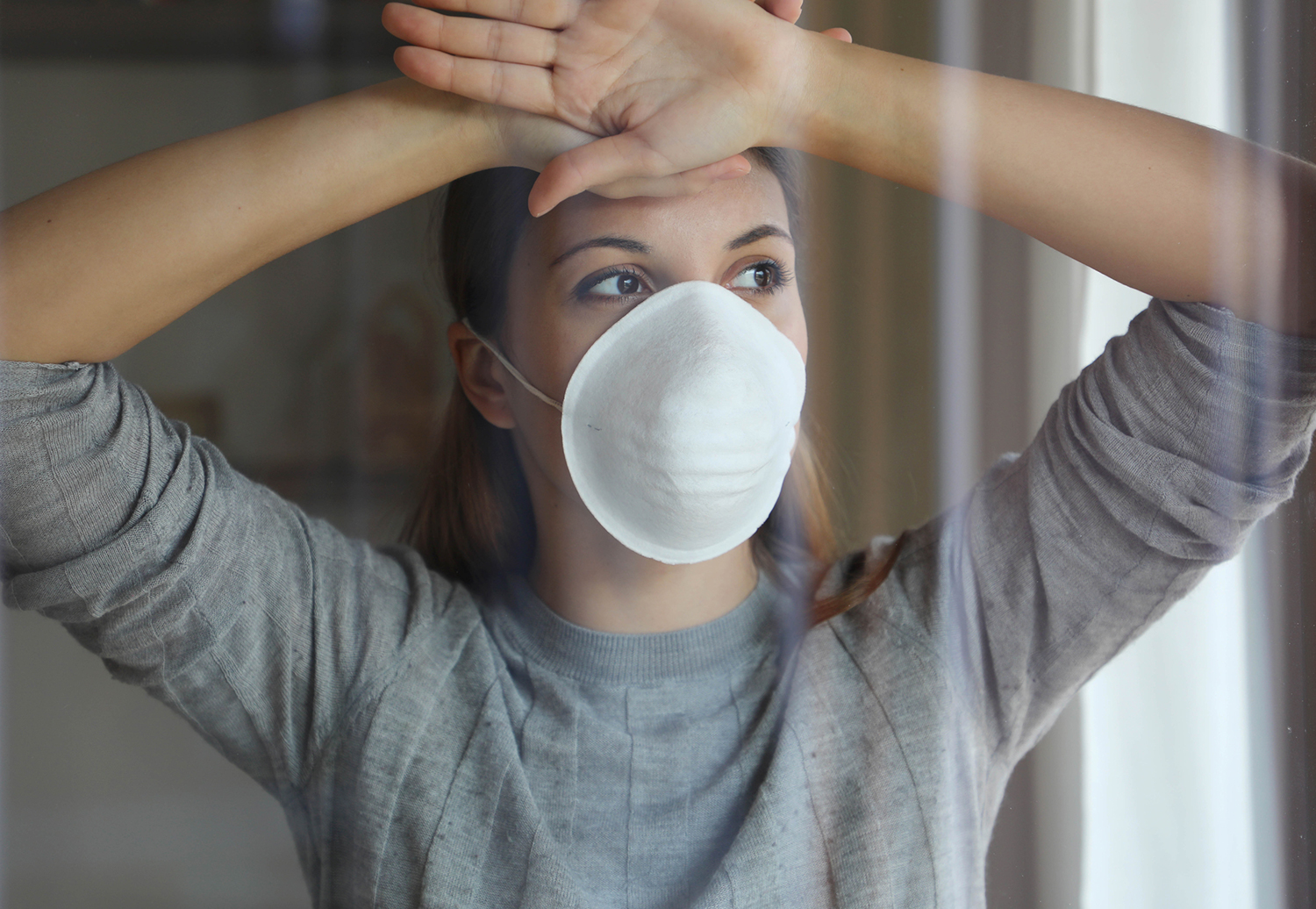 The Anxiety and Grief of COVID-19 A woman in a face mask looking longingly out of a window.