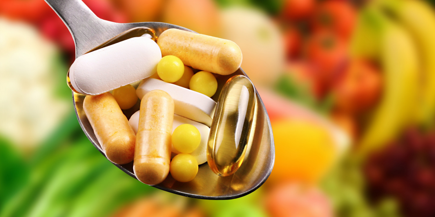 A spoon full of different supplements with fruits and vegetables in the background.
