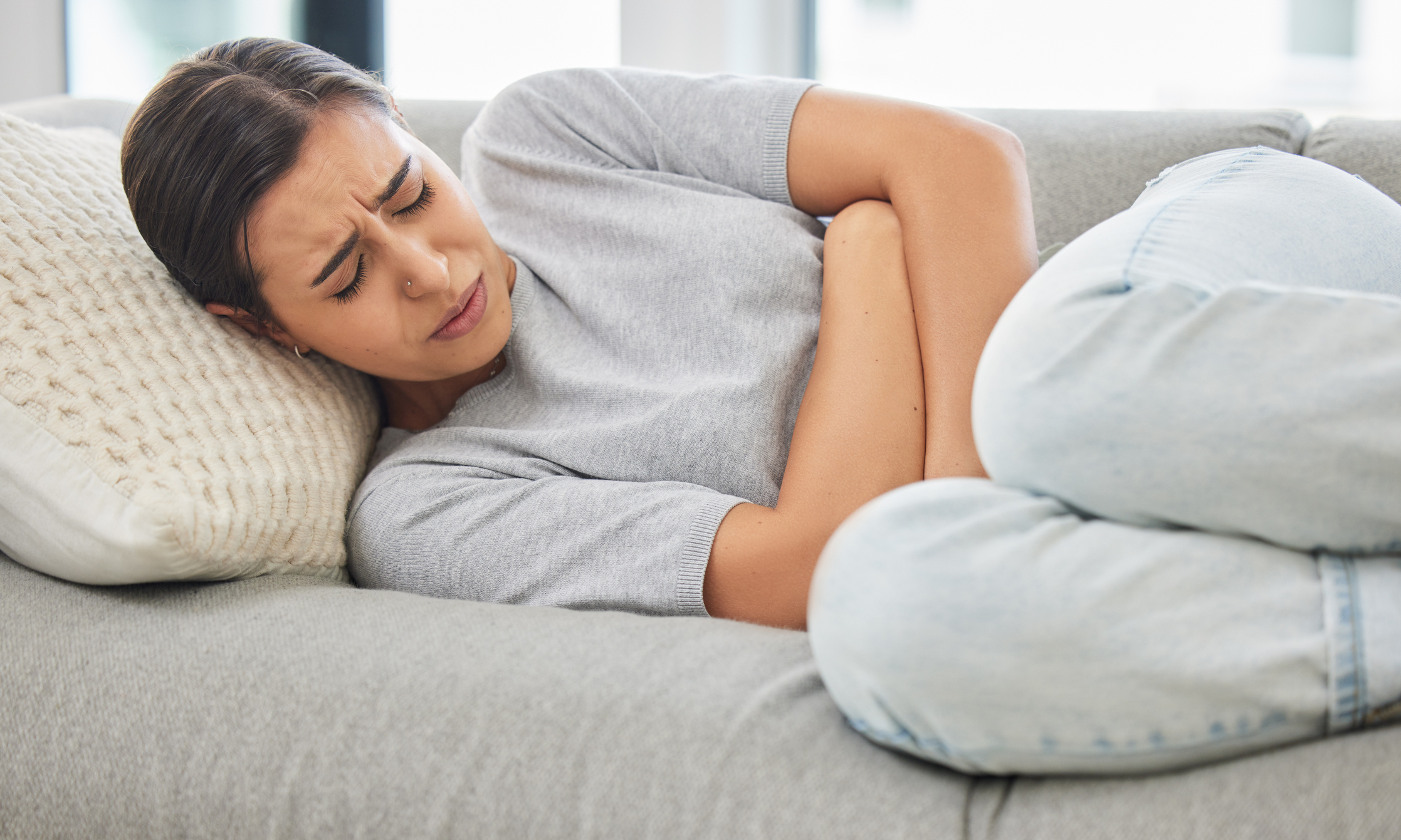 Young woman in pain with eyes closed curled into a ball on a grey couch.