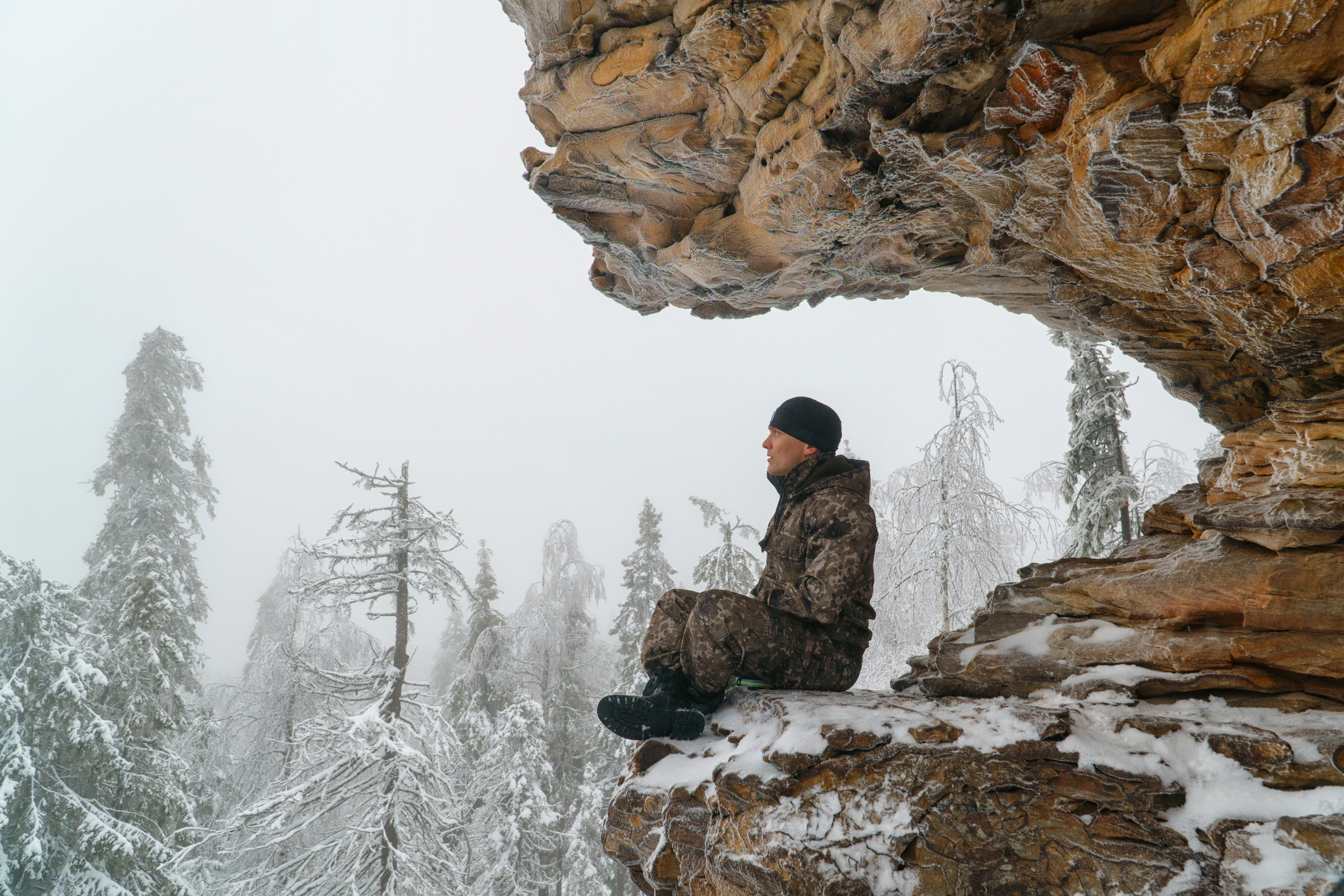 Man dressed in camouflage and a hat meditating on a mountain in winter. 