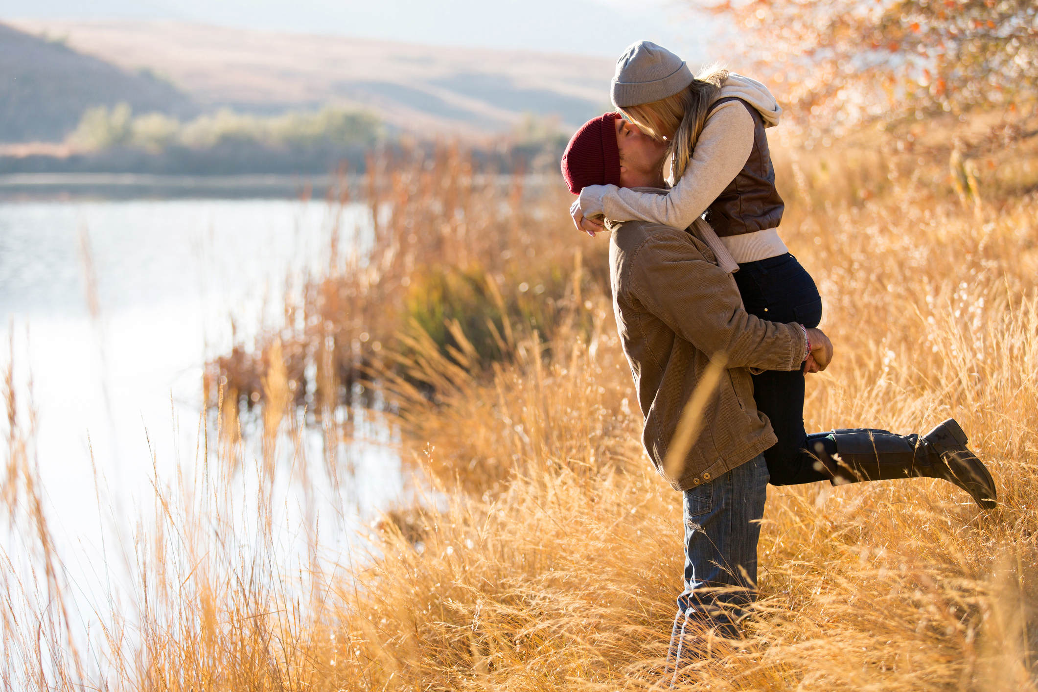 Man holding a woman in the air beside a mountain lake in autumn.