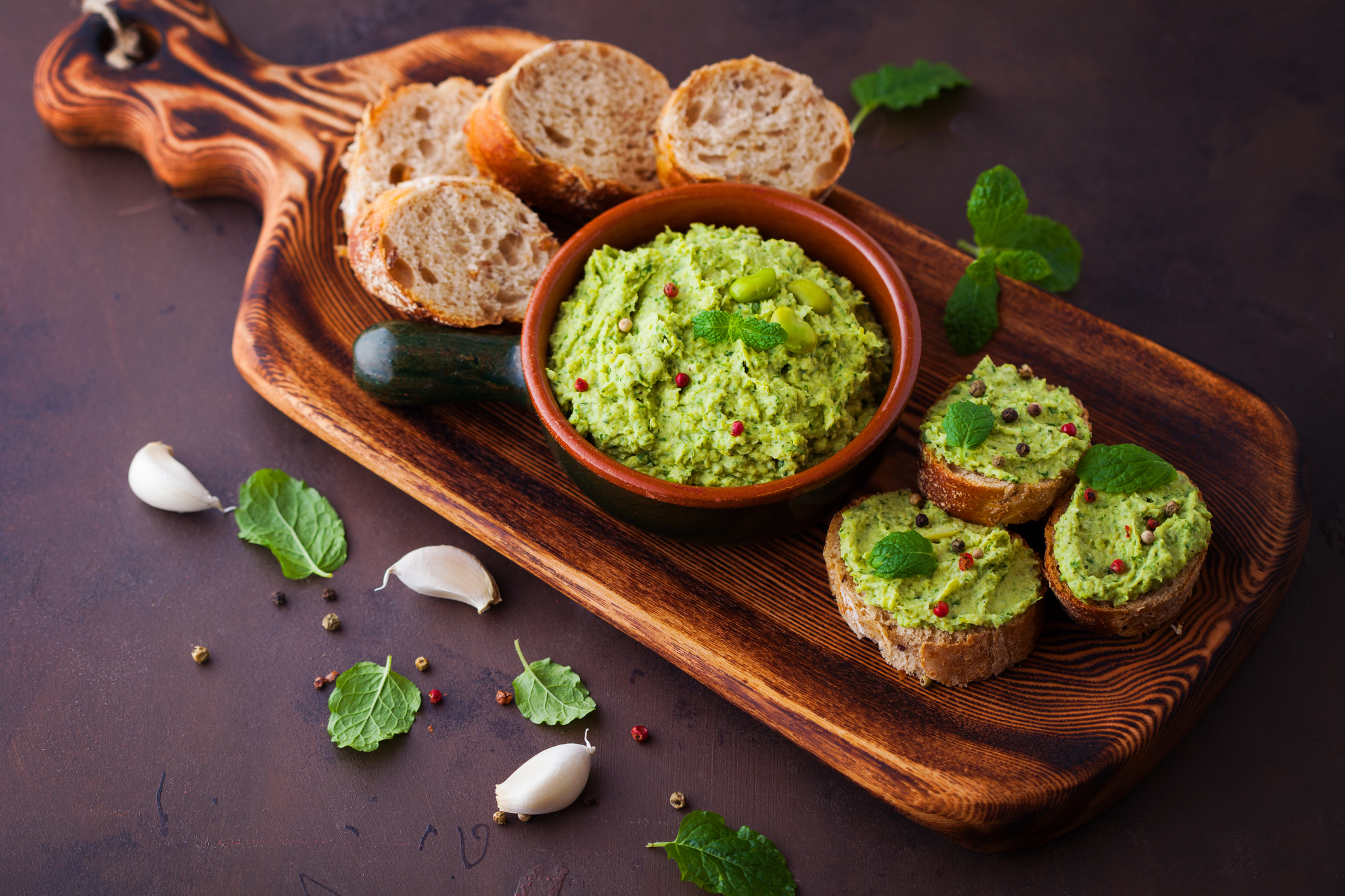 Fava Bean Hummus Bowl of fava hummus on a bread board along with bread rounds and three pieces of bread with fava hummus on them.