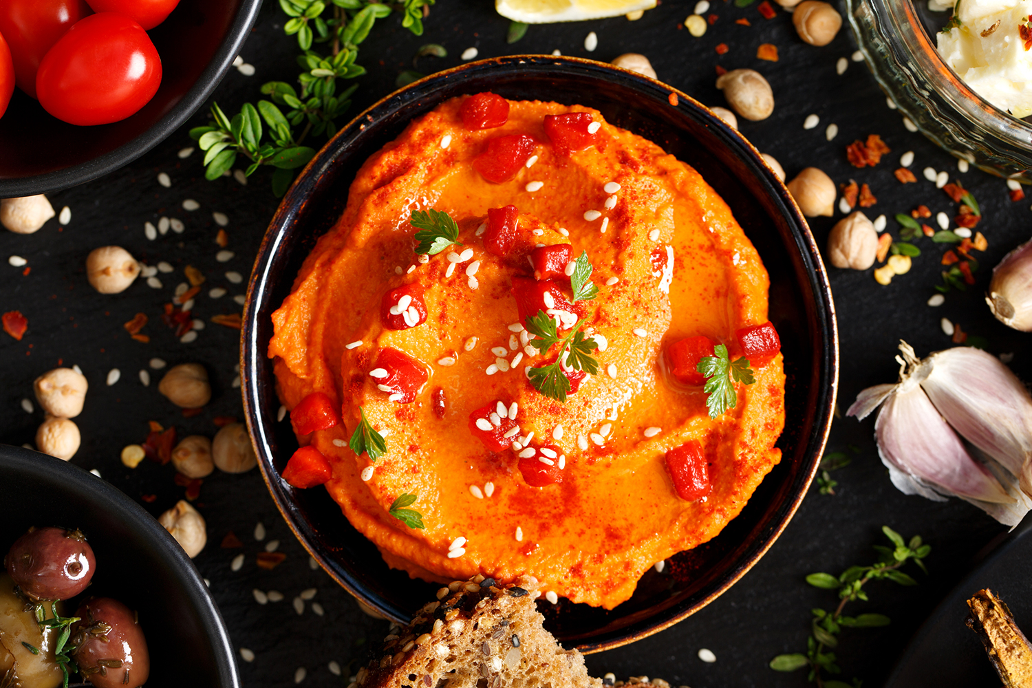 Top view of Red Hummus in a dark colored bowl surrounded by ingredients and foods to dip. Dark background.