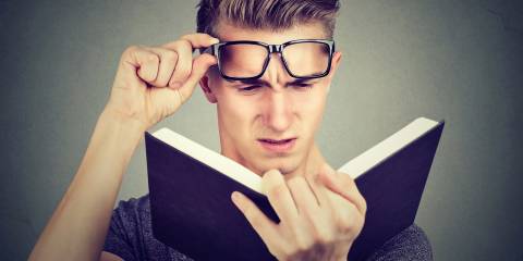 A young man lifting his glasses straining to read a book.
