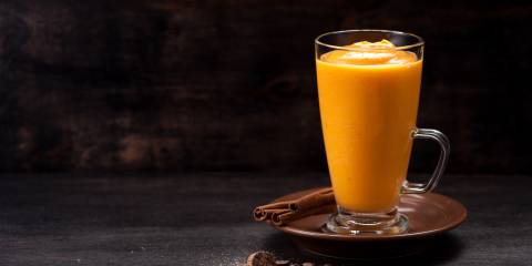 A glass cup filled with pumpkin spice smoothie next to cinnamon sticks. 