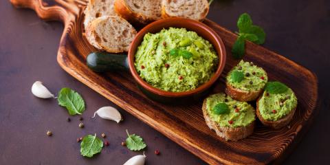 Bowl of fava hummus on a bread board along with bread rounds and three pieces of bread with fava hummus on them. 