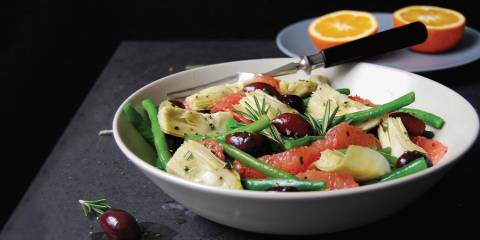 Green Bean, Artichoke, & Grapefruit Salad with Olives and Rosemary in a white bowl with a cut orange in the background. Black background.