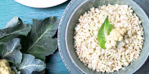 Raw Cauliflower Rice Cauliflower rice in a blue bowl on a blue table next to a head of cauliflower with leaves.